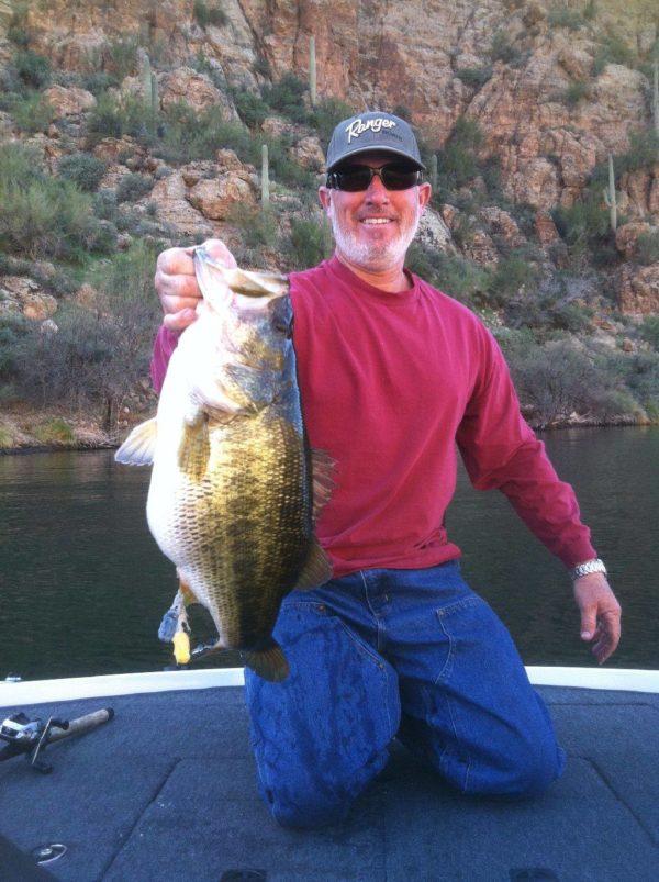 man in a red shirt on a boat on a lake holding up a humongous bass for the camera and smiling ear to ear
