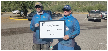Two anglers holding a weigh in board with their stats written on it in very illegible handwriting, sorry.